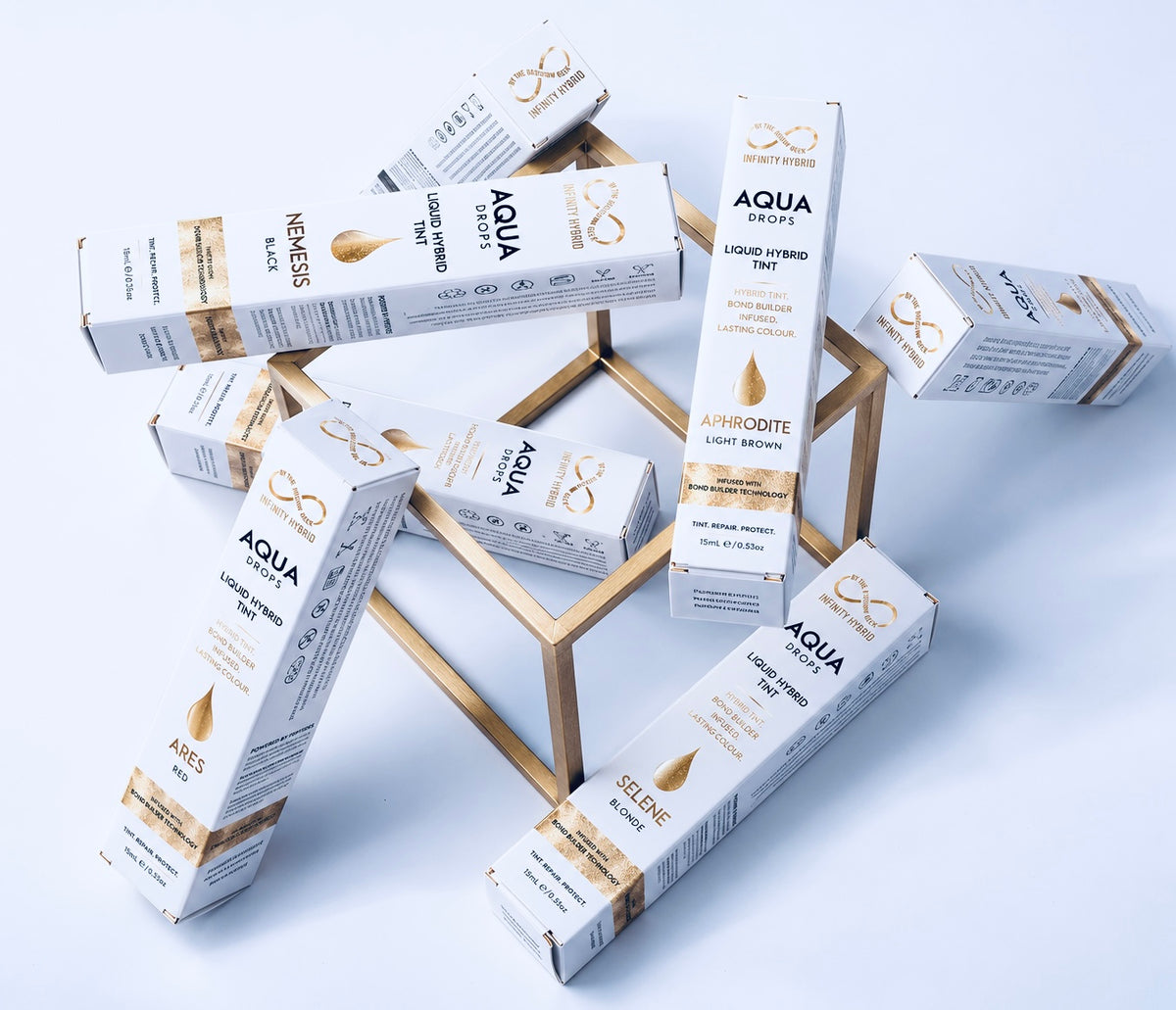 Aqua Drops® liquid hybrid brow dye 15ml boxes in Nemesis, Aphrodite, Ares and Selene shades on a gold stand, Infinity by The Brow Geek hybrid brow tint